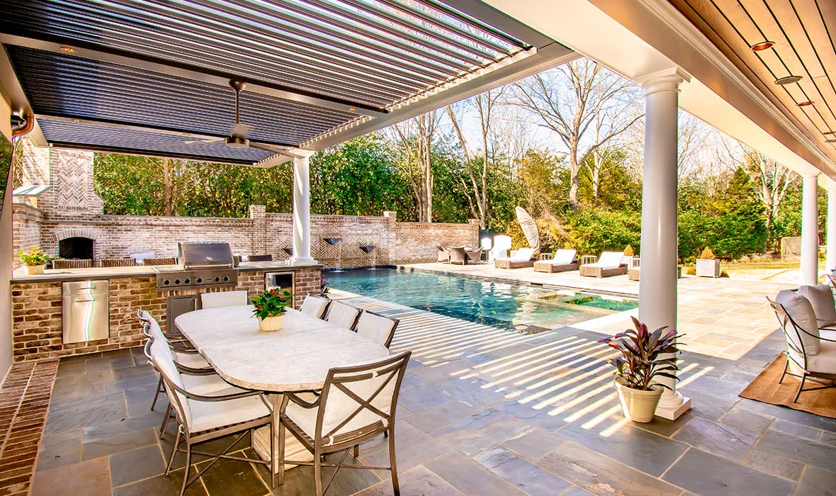 An immaculate poolside patio includes a shaded dining area, barbecue, and bar.