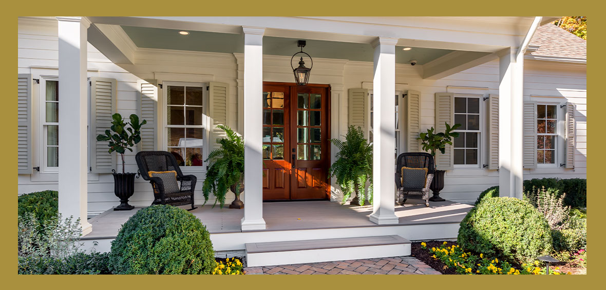 A front porch features a couple of chairs and potted trees with a large double-door in the center and manicured landscaping up to the steps.