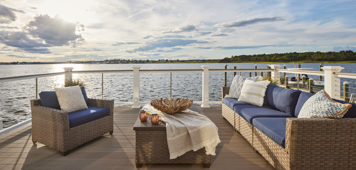 A lakeside deck with glass railing has a matching furniture set including an outdoor sofa, coffee table, and armchair. 