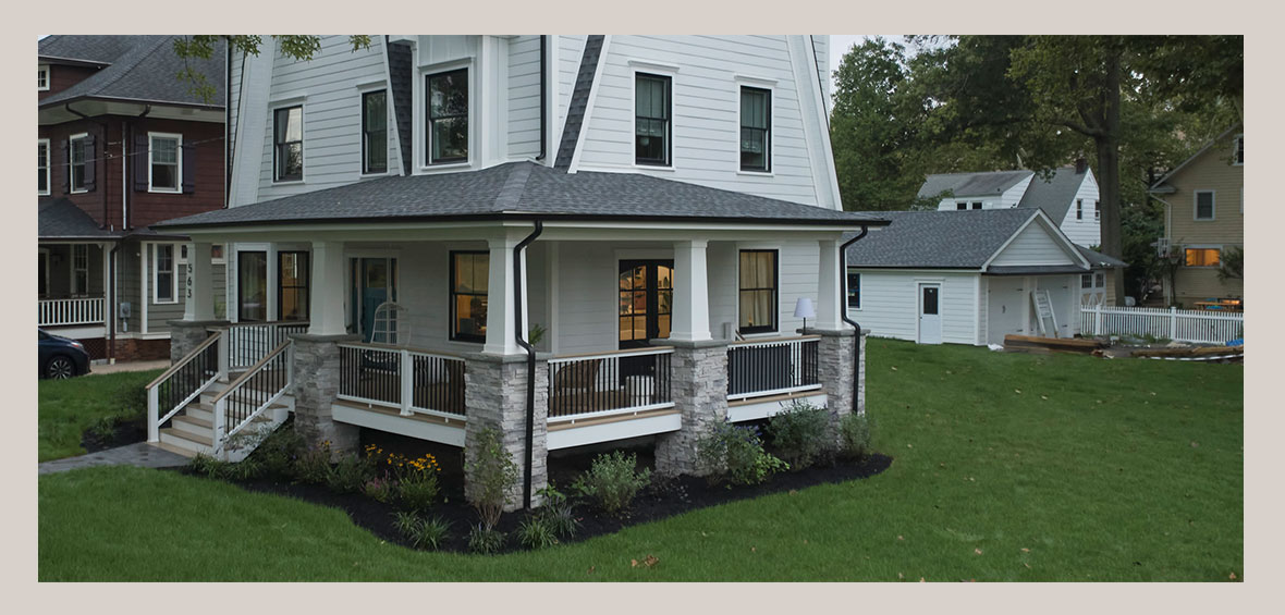  A double-story home features a roofed veranda wrapping around two sides of the home with black railing and a flower bed around the perimeter.