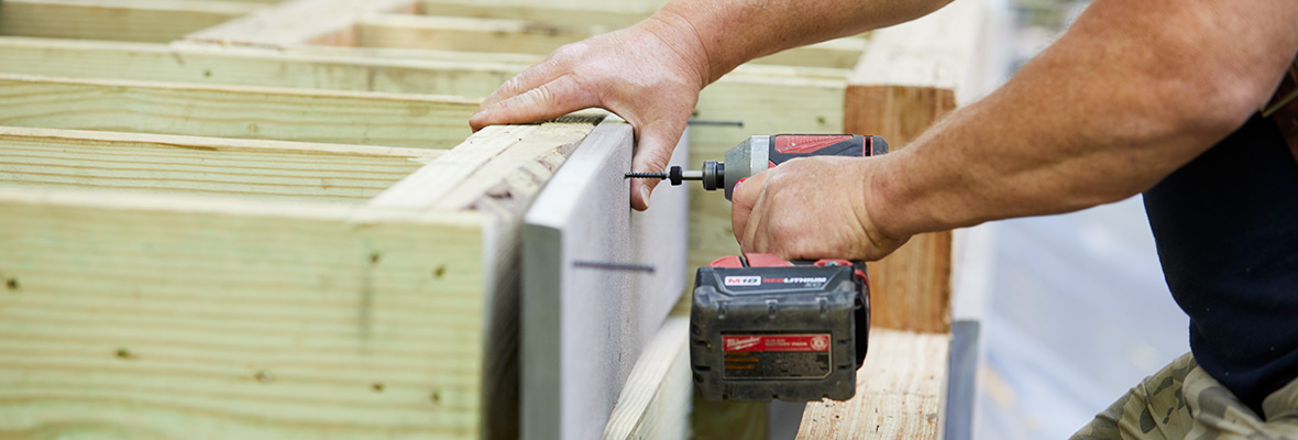 A contractor uses a power drill to fasten a decking rim board to a rim joist while installing a deck.