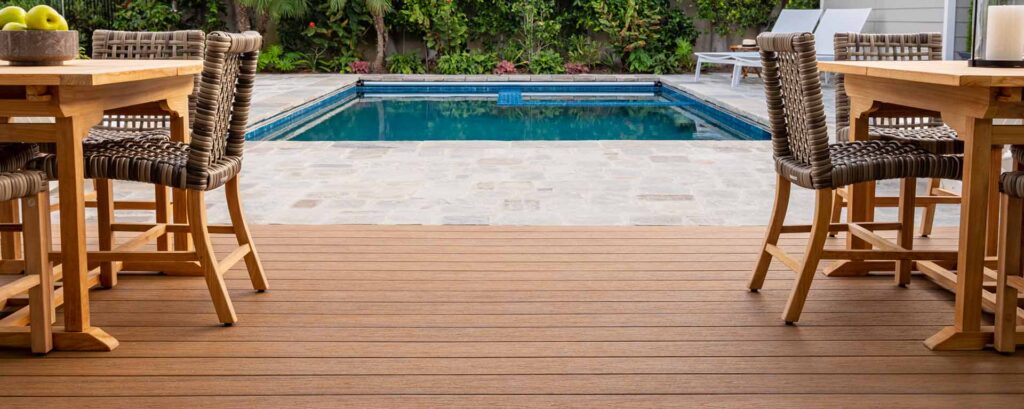 Two table and chair sets sit upon a warm brown deck with a sunken pool in the background