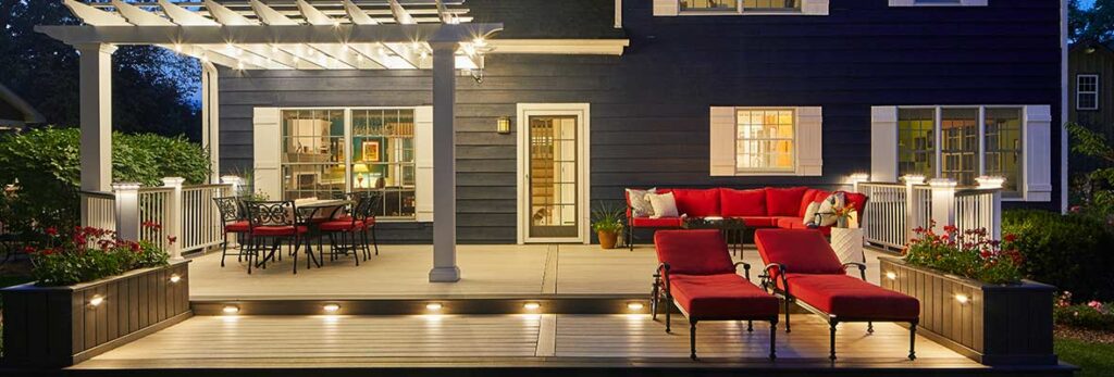 A backyard deck features built-in lighting in steps and post caps, with a pergola wrapped in string lights above a dining set.