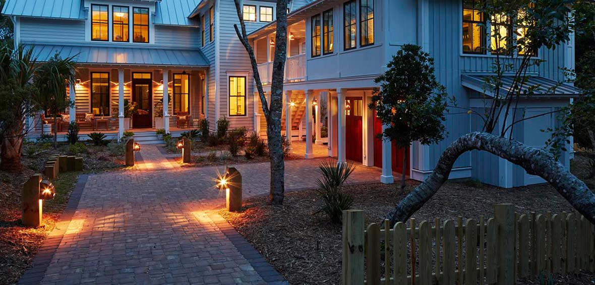 Interior lights, wall fixtures, and path lights add a warm yellow glow at dusk around a home and yard. 