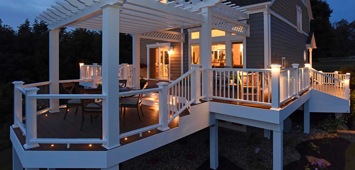 A stilted deck wraps around a home with built in deck and railing lights illuminating a pergola covered dining platform.