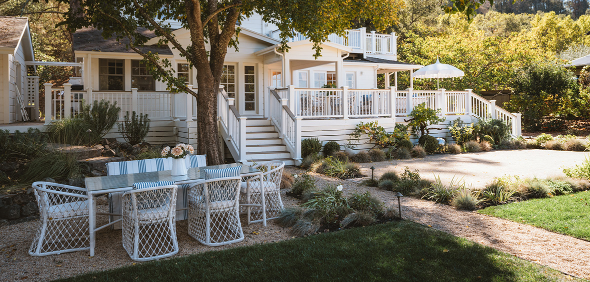 A long dining table sites under a tree on a gravel patio, surrounded by landscaping, garden paths, and lights.