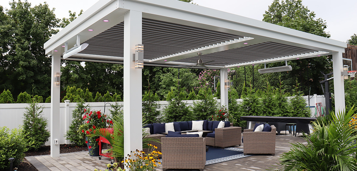A pergola with lights and a ceiling fan provides shade for an outdoor seating area surrounded by palms, trees, and potted flowers. 