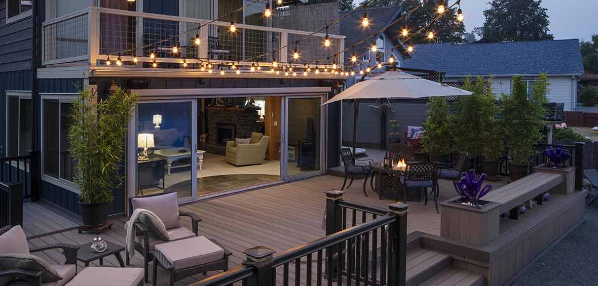 String lights stretch over a deck at dusk with a fire pit and lounge chairs below.