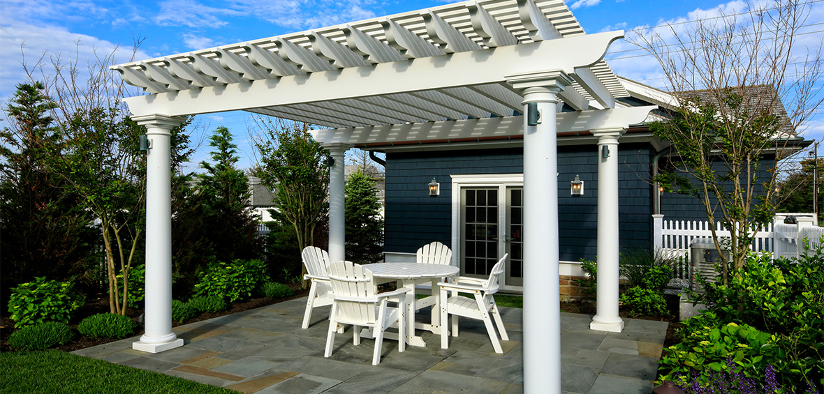 A white pergola protects a patio and dining furniture with shrubs and flowers surrounding the space. 