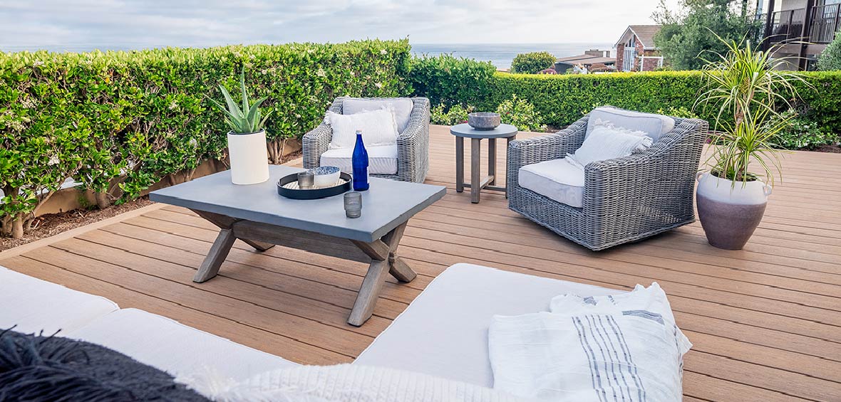 Two gray upholstered armchairs are placed in an outdoor deck seating area surrounded by tall hedges and potted plants. 