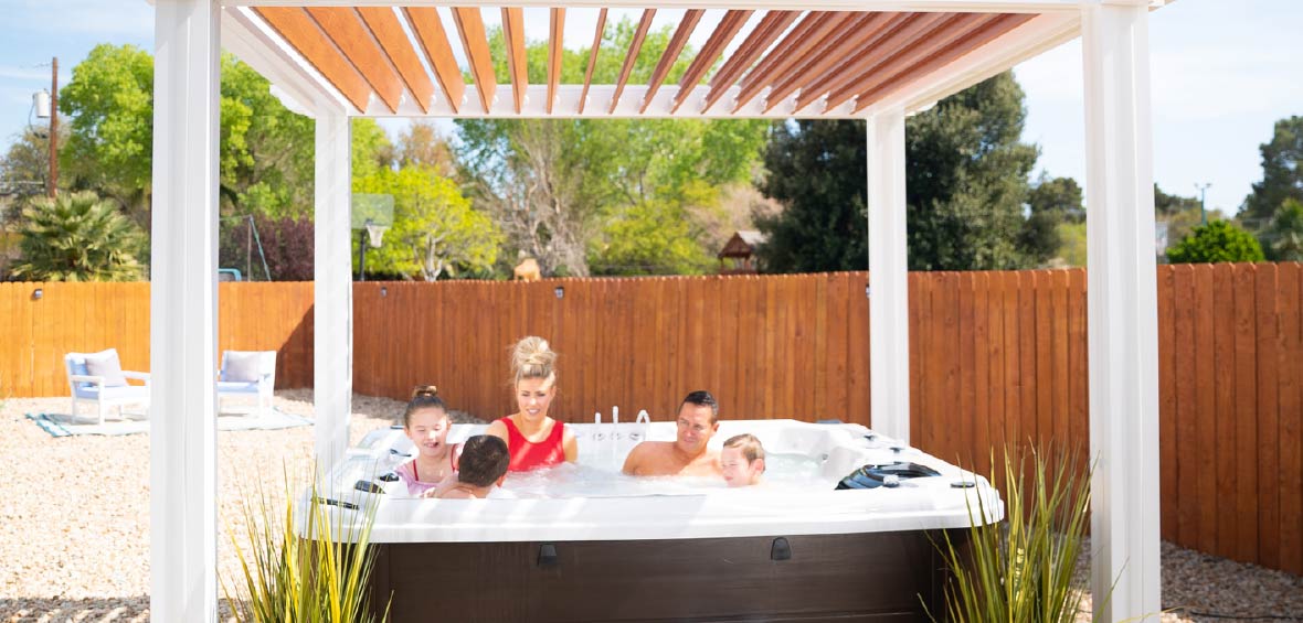 A family enjoys time in a personal hot tub surrounded by a privacy fence with a pergola overhead.