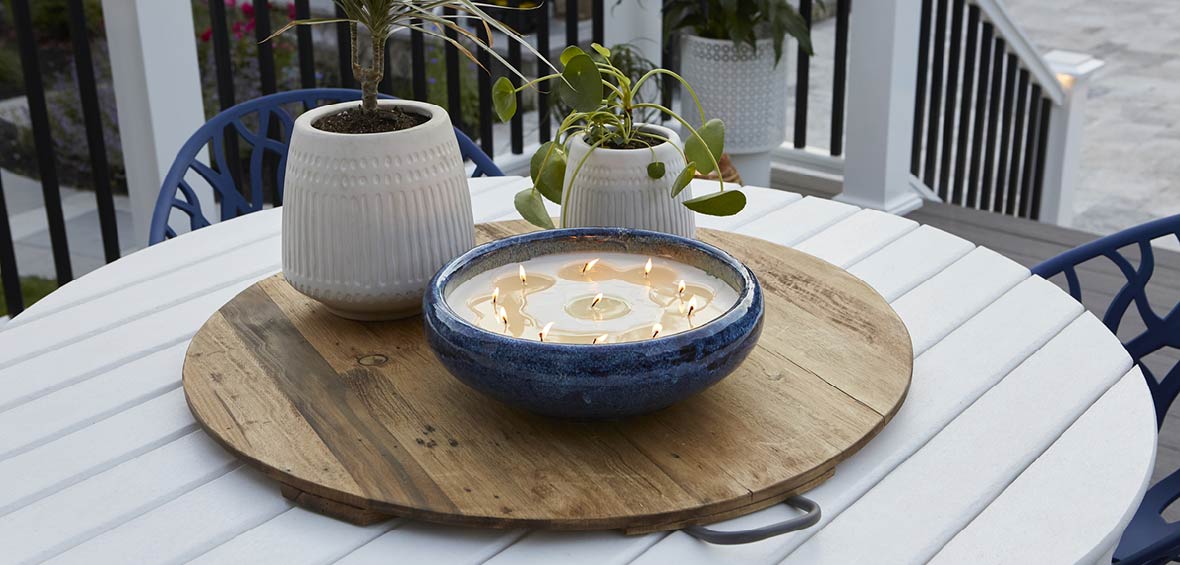 Two potted plants sit next to a large outdoor candle as part of a centerpiece on a deck table. 