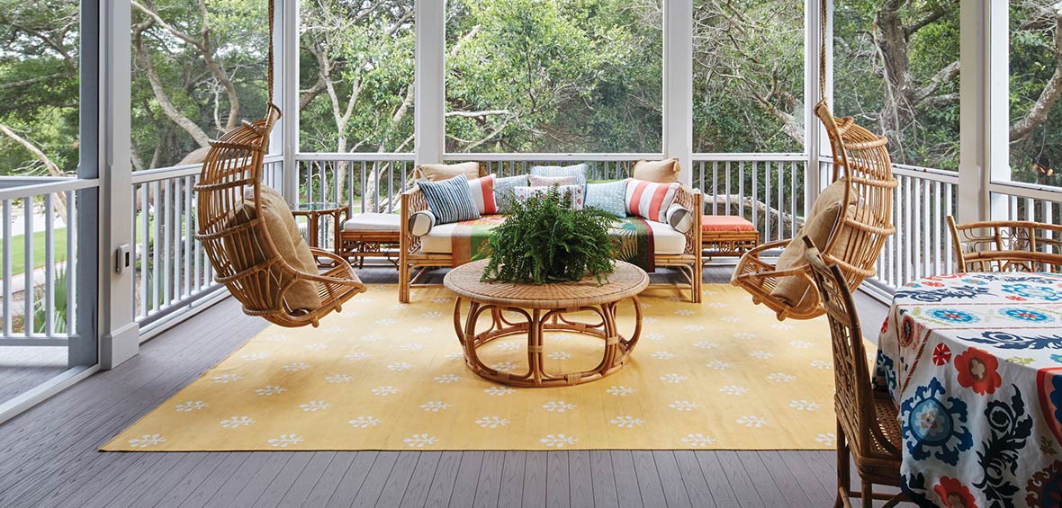 Rattan deck furniture features plush pillows, bold patterns, and potted plants for a cozy aesthetic. 