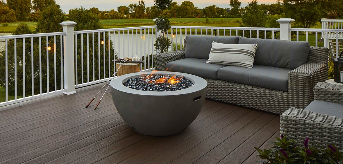 A lit fire pit is centered in a deck’s outdoor living area with skewers and s’more supplies on a nearby table.