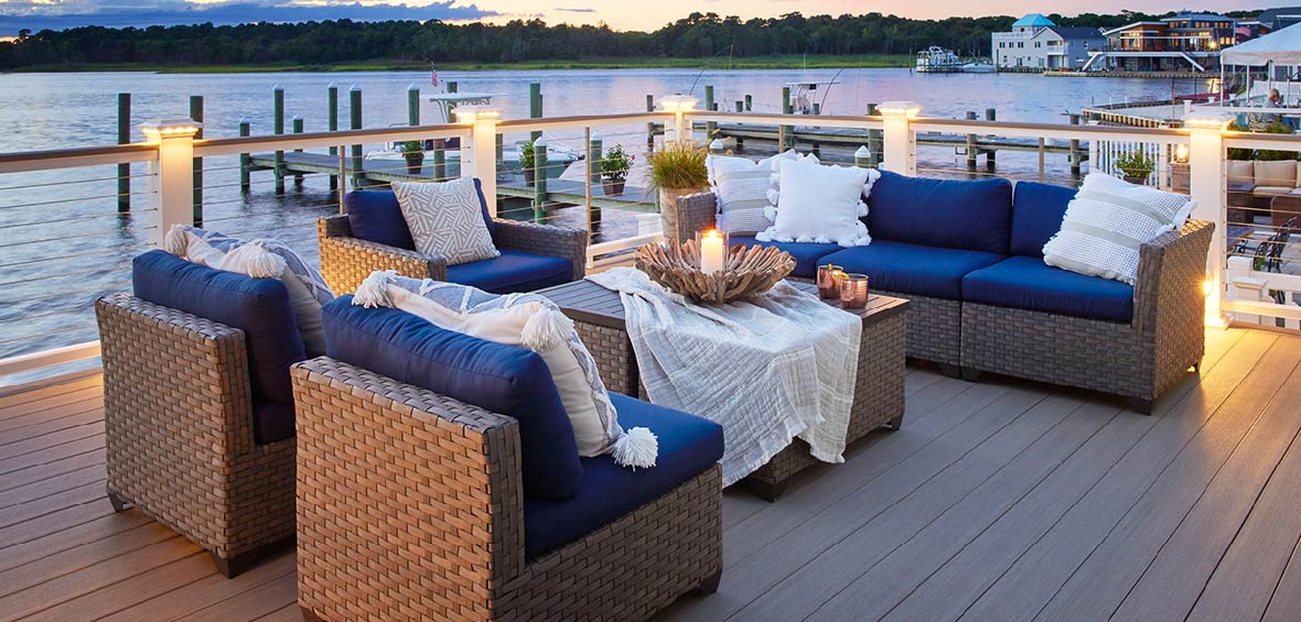 An outdoor living area with seating and tables sits on a harbor deck with post cap lights providing a gentle glow at dusk.