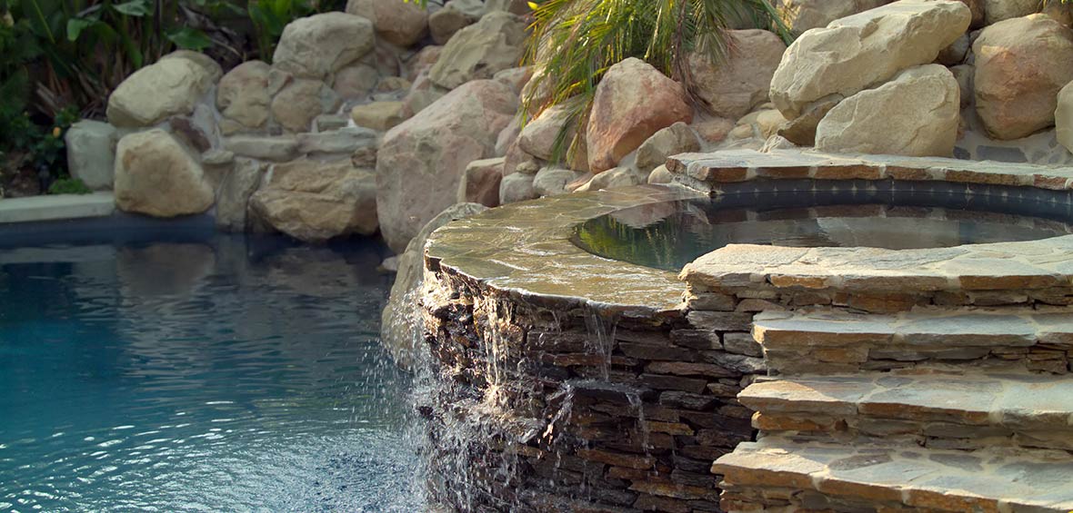 A stone hot tub doubles as an overflow fountain into an adjacent pool, and both features are surrounded by stones. 
