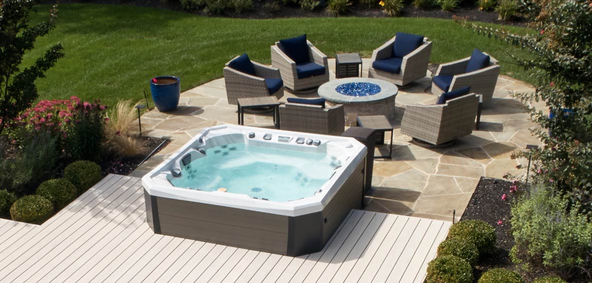A hot tub at the edge of a deck and patio is surrounded by landscaping and blocked by a seating area and fire pit.