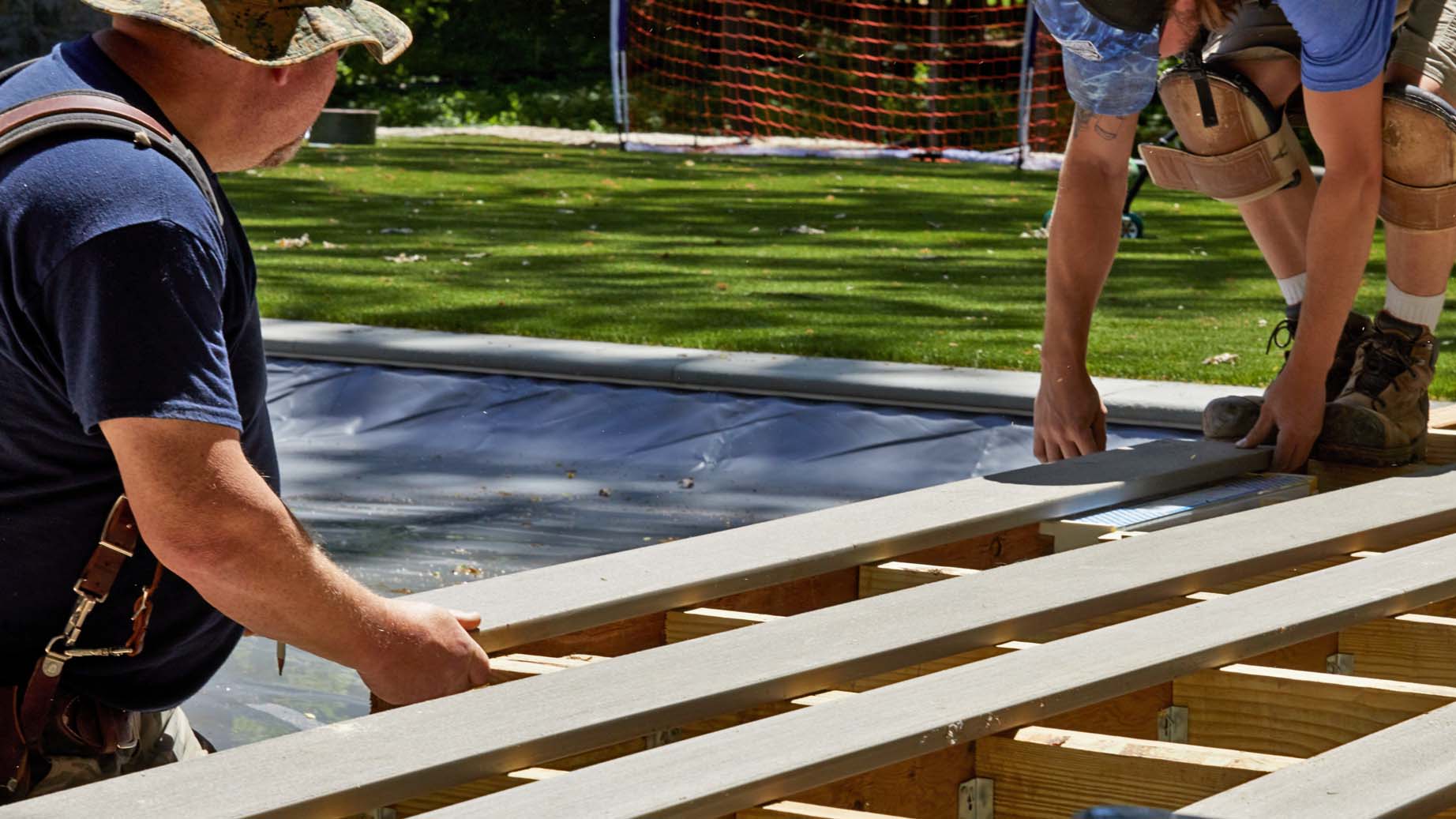 Two men on a construction site measure light tan colored deck boards