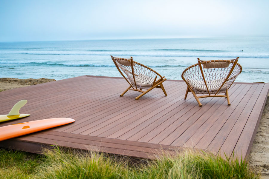 Brown deck with a reddish hue on the beach with two chairs and two surf boards laying on top of it