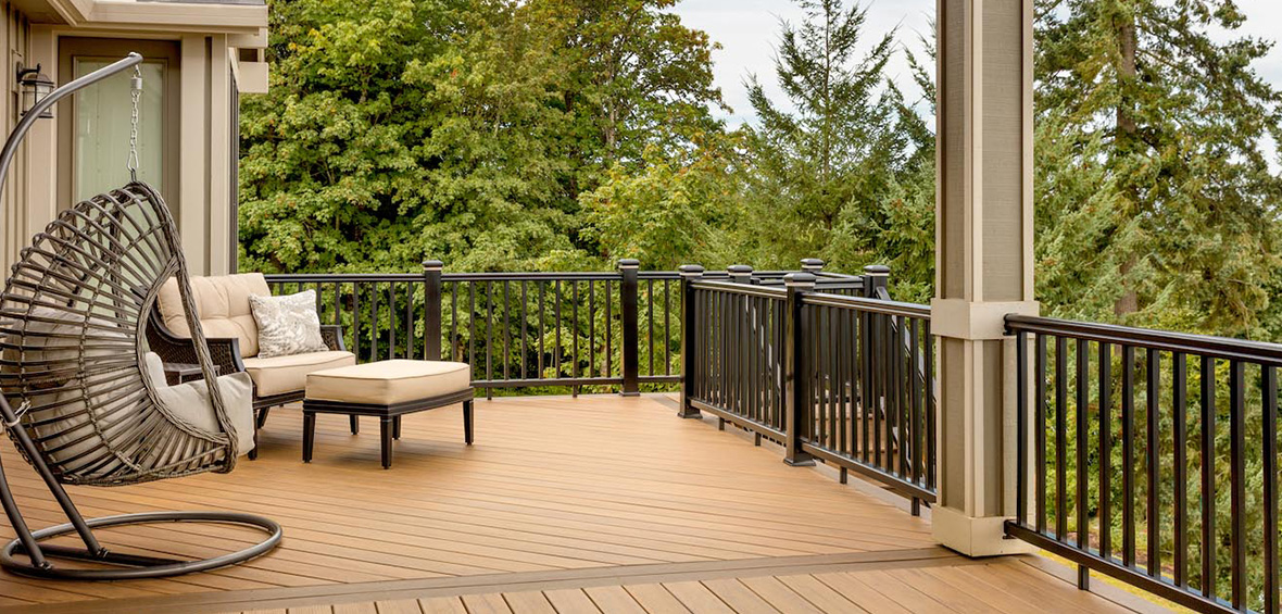 An elevated deck features an armchair, ottoman, and a suspended chair looking out over pine trees with a set of stairs to the yard.