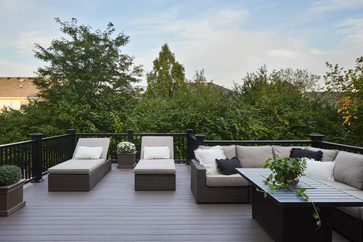 A sectional couch, coffee table, and two padded lounge chairs sit on a second story deck surrounded by trees