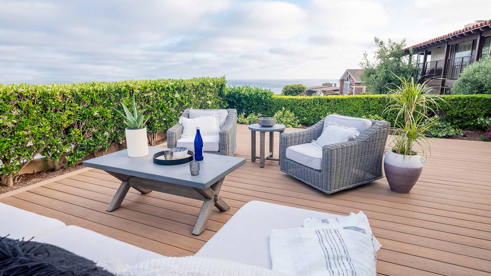 Gray, wicker outdoor armchairs on a reddish-brown deck surrounded by tall hedges