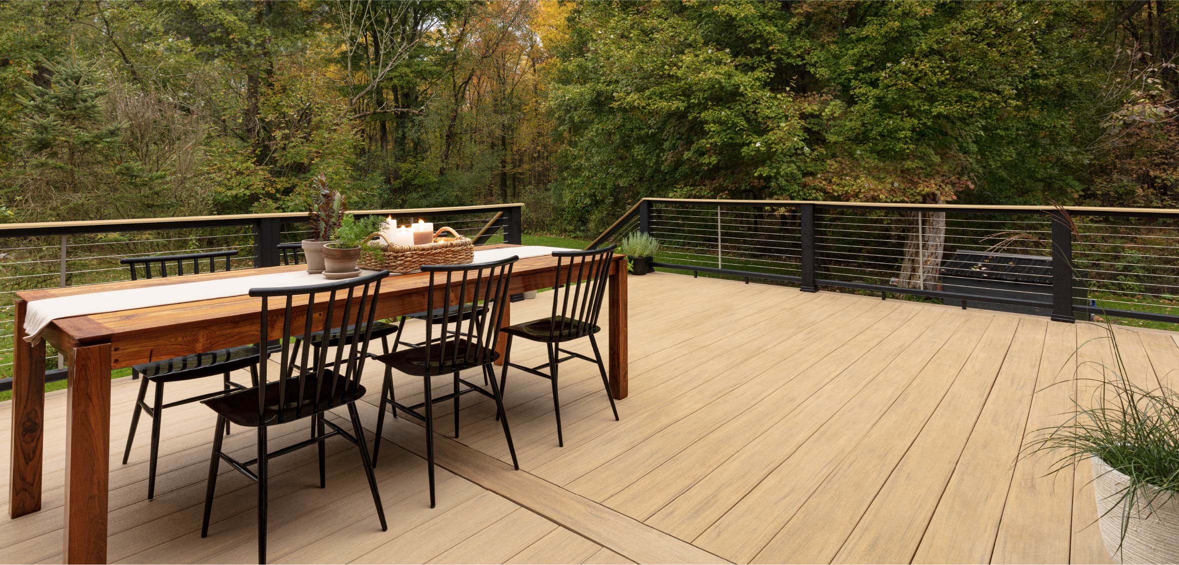 A long dining table with six chairs is set on a light wood deck with black cable railing along the perimeter.