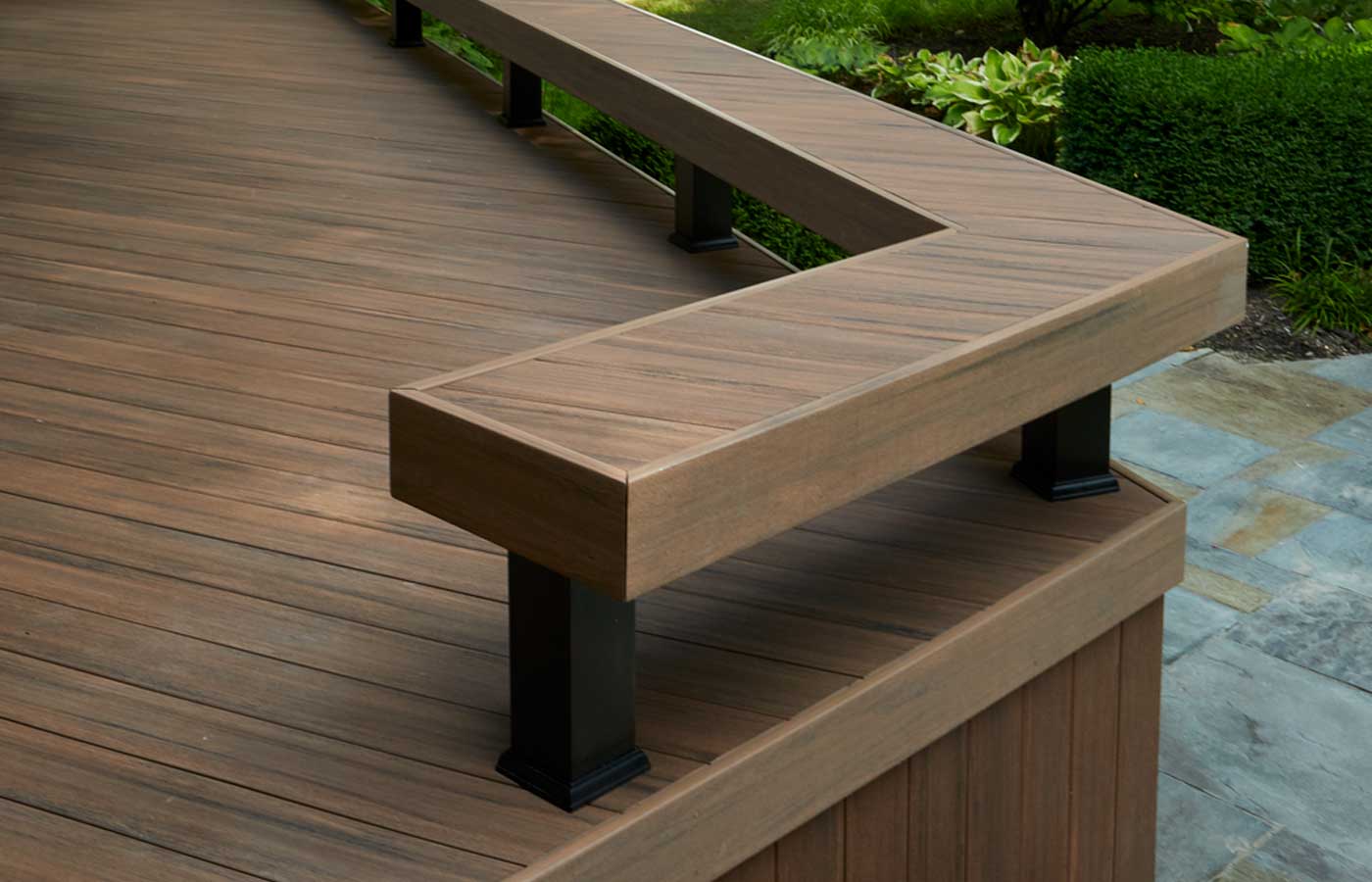 A close-up shot of a composite deck with diagonally laid deck boards. A built-in bench runs along the deck’s perimeter. Greenery surrounds the deck, suggesting it's built in a wooded area. The composite deck is made of English Walnut boards from the TimberTech Advanced PVC Vintage Collection.