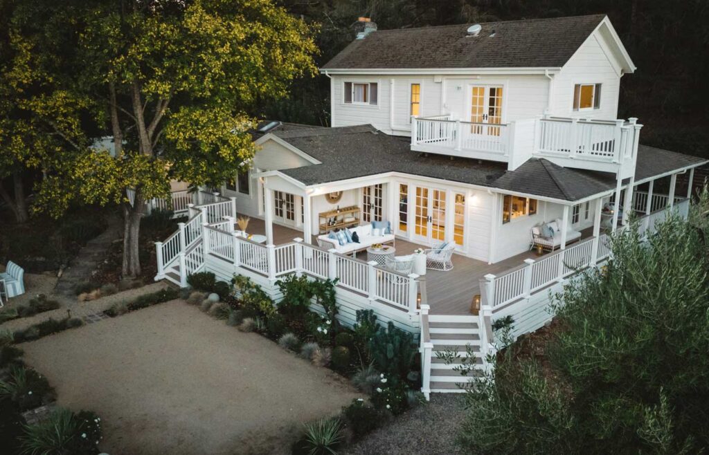 A large, white home surrounded by trees and tall bushes has windows illuminated in the early eve