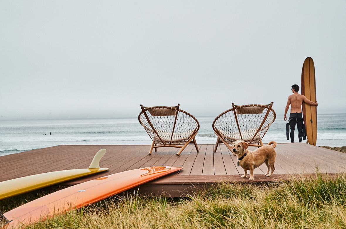 A floating deck next to the beach features two chairs and surfboards, with a dog and surfer standing nearby.
