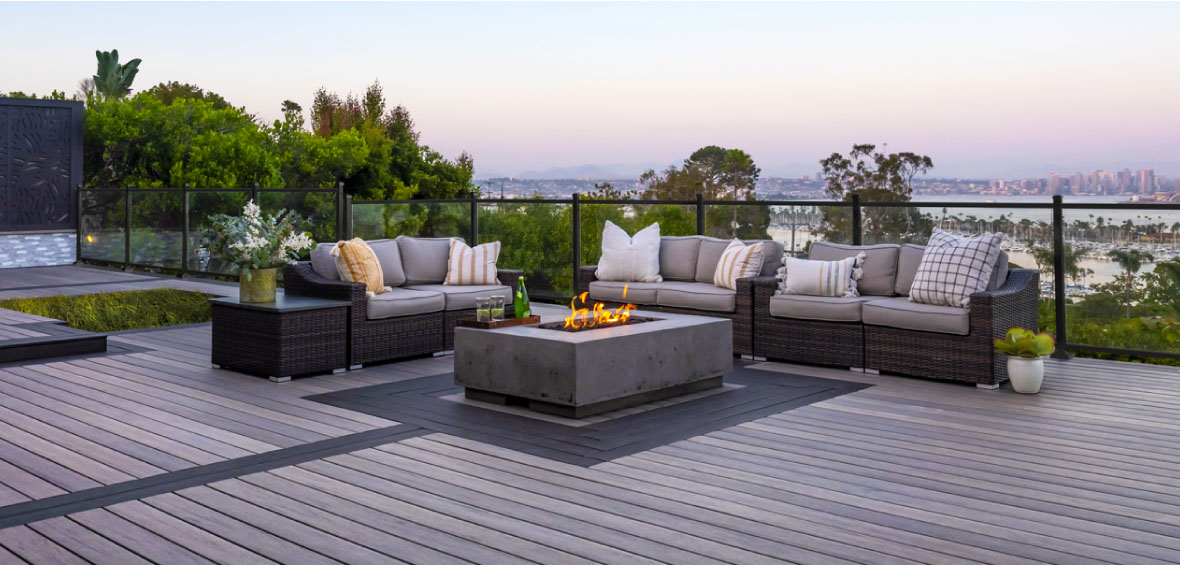 A three-piece gray seating set faces a centered firepit on a deck inlay. The deck is gray with dark contrasting boards. 