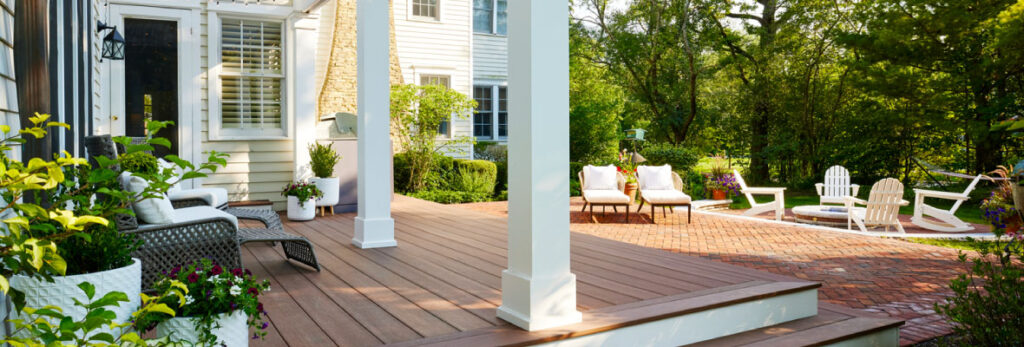 A brown deck connects a backyard pool to a home with white columns, lounge furniture, and landscaping.