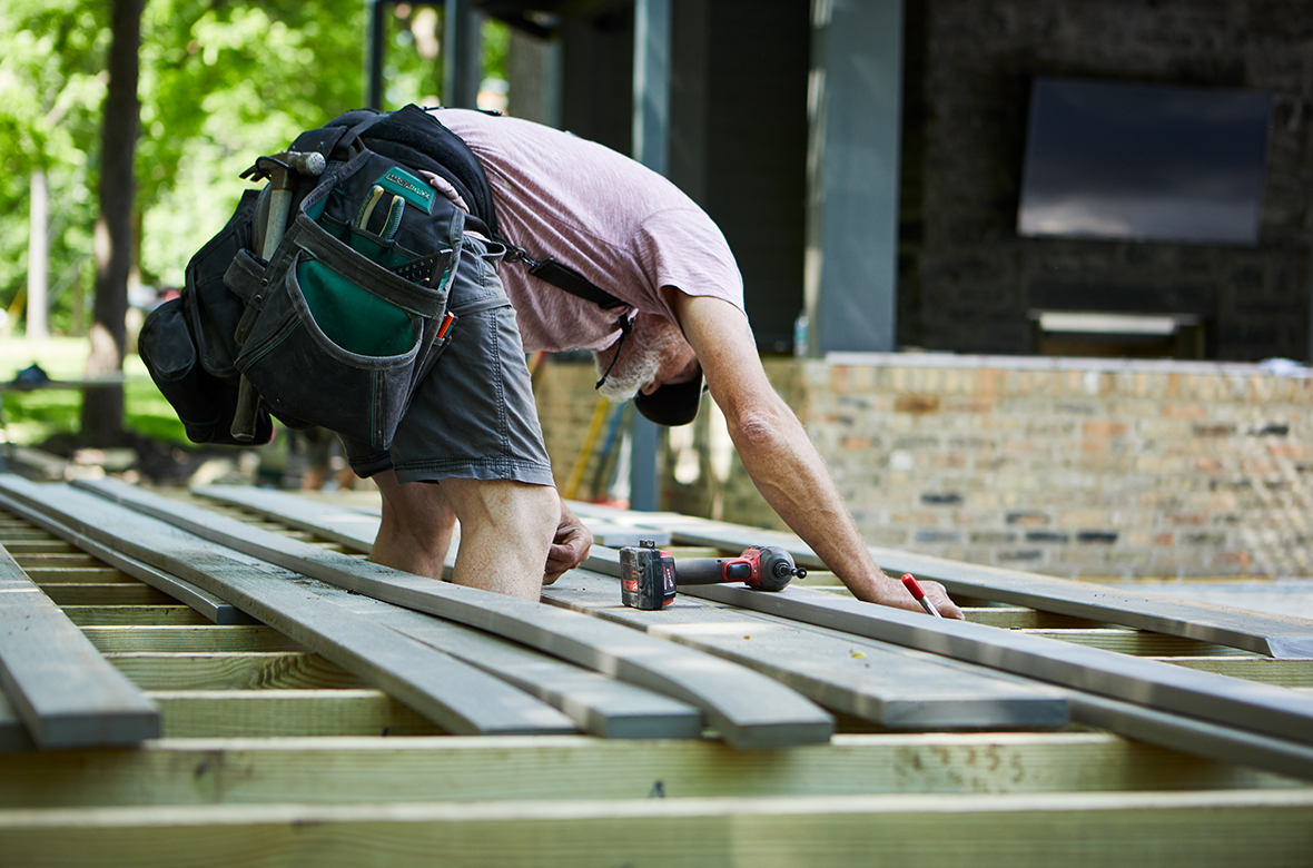 A builder leans over for a closer look at a deck’s substructure, with decking and a power drill laying around him.