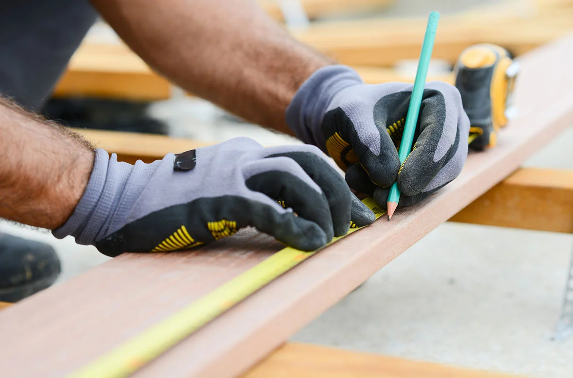 A close up of gloved hands holding a measuring tape flat and marking a measurement on a deck board. 