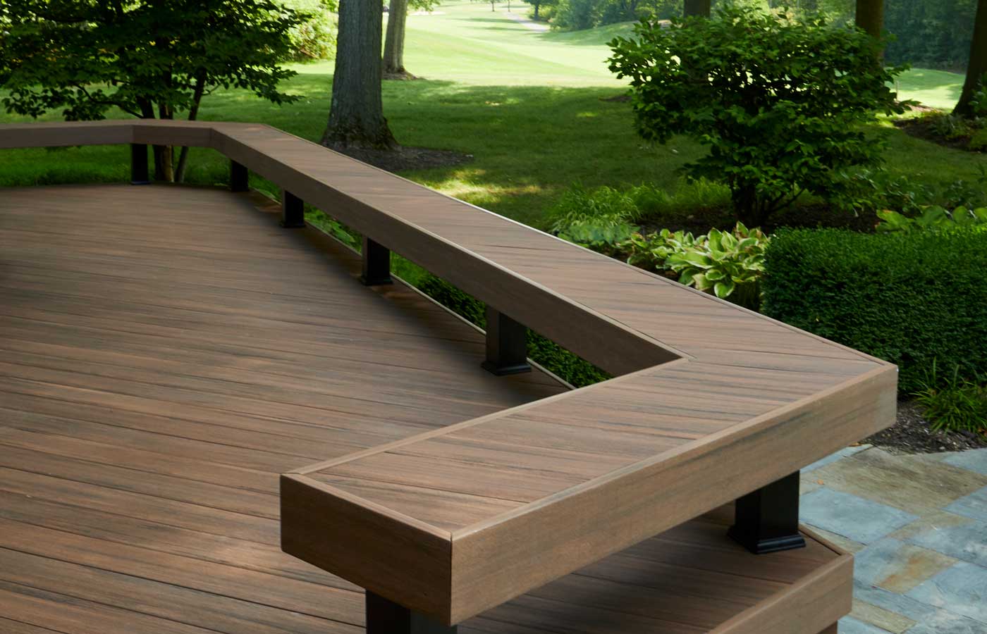 A close-up shot of a composite deck with diagonally laid deck boards. A built-in bench runs along the deck’s perimeter. Greenery surrounds the deck, suggesting it's built in a wooded area. The composite deck is made of English Walnut boards from the TimberTech Advanced PVC Vintage Collection.