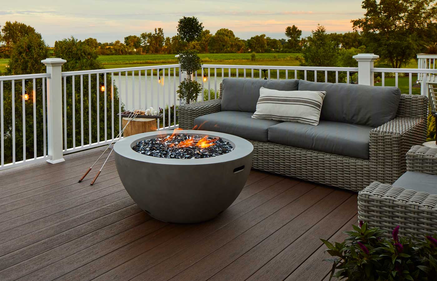 A dark brown composite deck with a white railing looks out at the last gasp of the sunset, firepit already lit