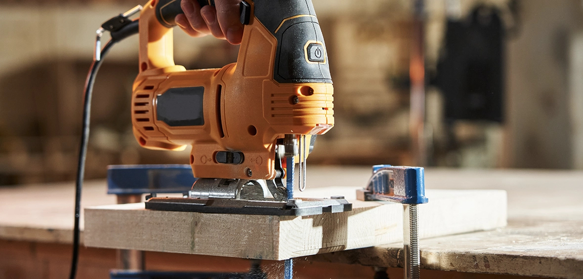 A handheld jigsaw features a narrow, vertical blade for precision cuts and is stopped in the middle of a cut on clamped lumber. 