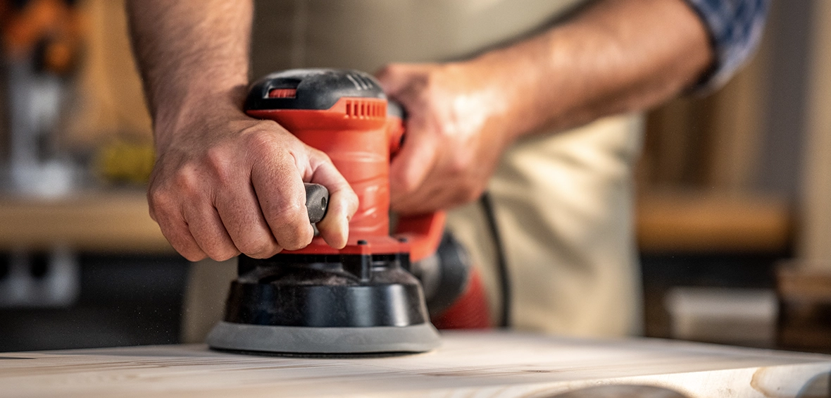 Someone holds and orbital sander and moves it across a wood surface to create a smooth finish. 