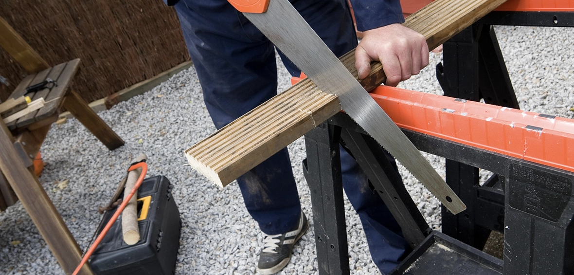 Someone uses a hand saw to cut a plank of wood positioned on two orange and black sawhorses. 