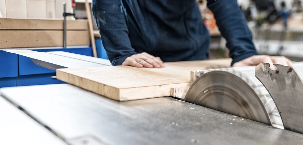 A saw blade comes up through the table of a table saw and is rotating as a carpenter pushes wood towards it. 