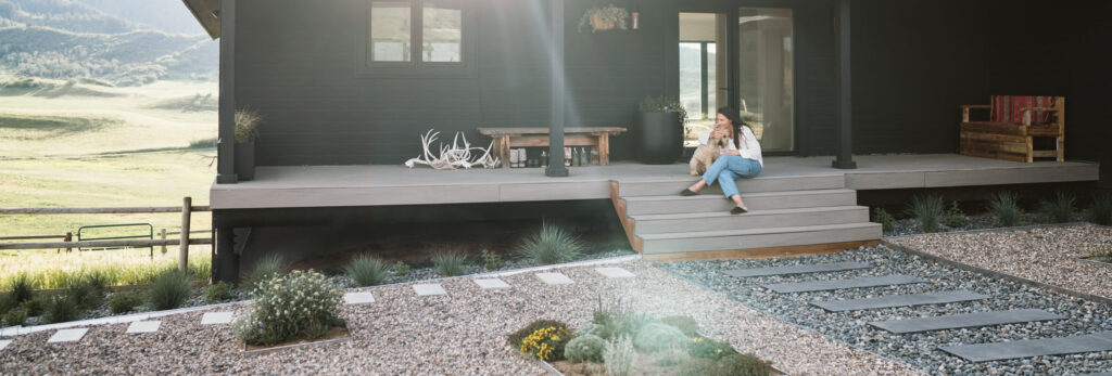 A woman and her dog sit on the steps of a deck with a sloping hill.