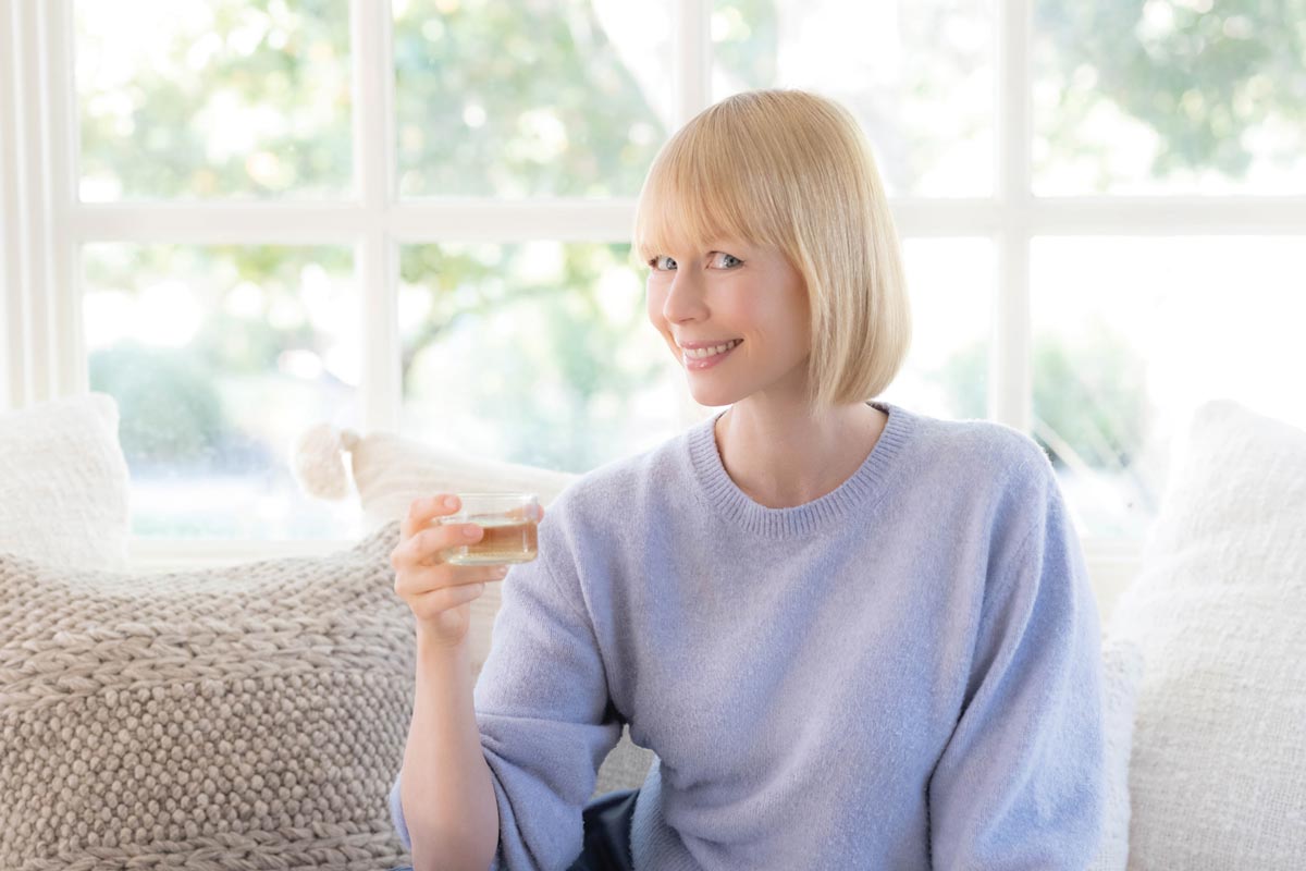 Erin poses for a portrait in her home. She has a blonde bob haircut and is wearing a baby blue colored sweater. She holds a small glass of wine.