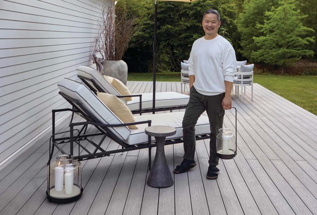 Danny Seo holds an elegant outdoor lantern and poses for a photo on his new TimberTech deck. The light gray deck has several lounge chairs on it and was built with Slate Gray deck boards from the Advanced PVC Harvest Collection
