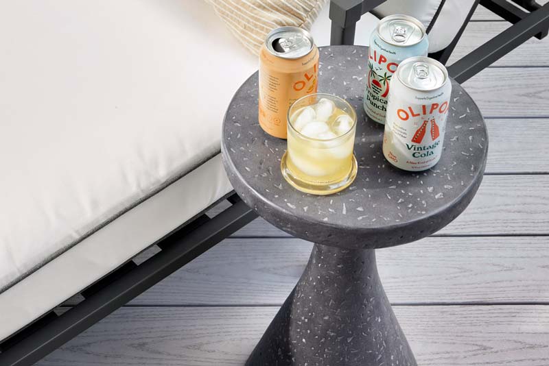 Cans of Olipop Vintage Cola and a rocks glass filled with a chilled beverage are shown atop a dark gray marble side table