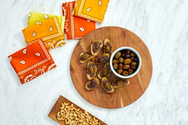 A platter of crostini breads with tapenade and a bowl of olives are on a marble table with brightly colored red, yellow, and orange bandanas