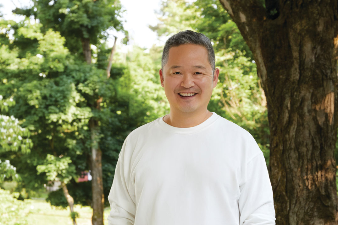 Headshot of Danny Seo, smiling and wearing a white sweater in his wooded backyard