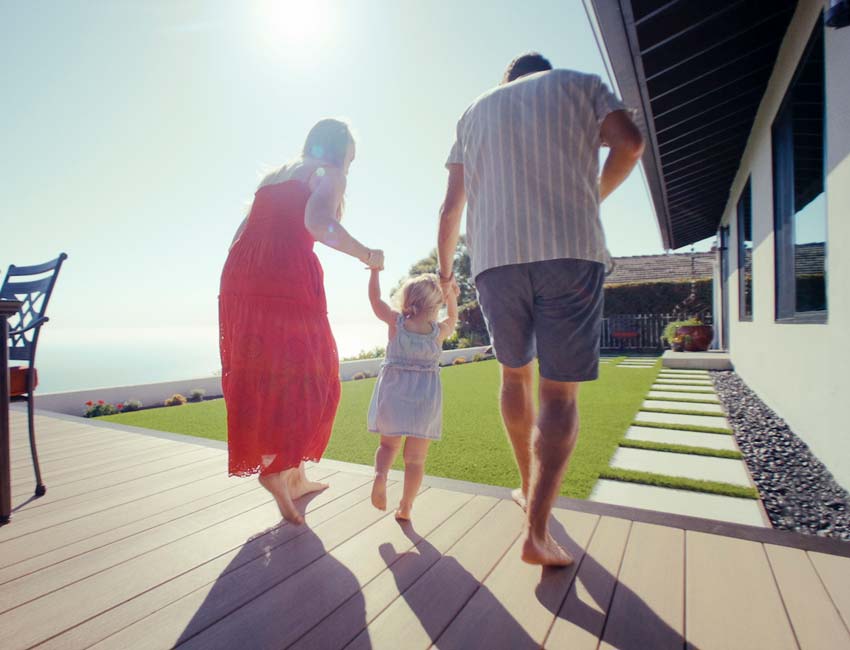 Mark and Jennifer's family members hold hands with a toddler as the trio walks barefoot on the deck towards the grass