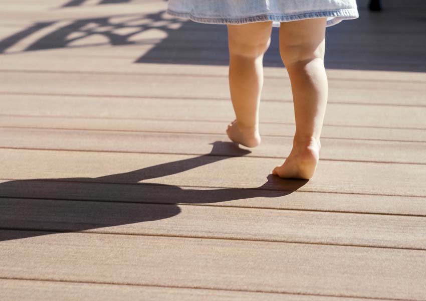 The legs and feet of a toddler in a denim dress are shown walking barefoot on a deck
