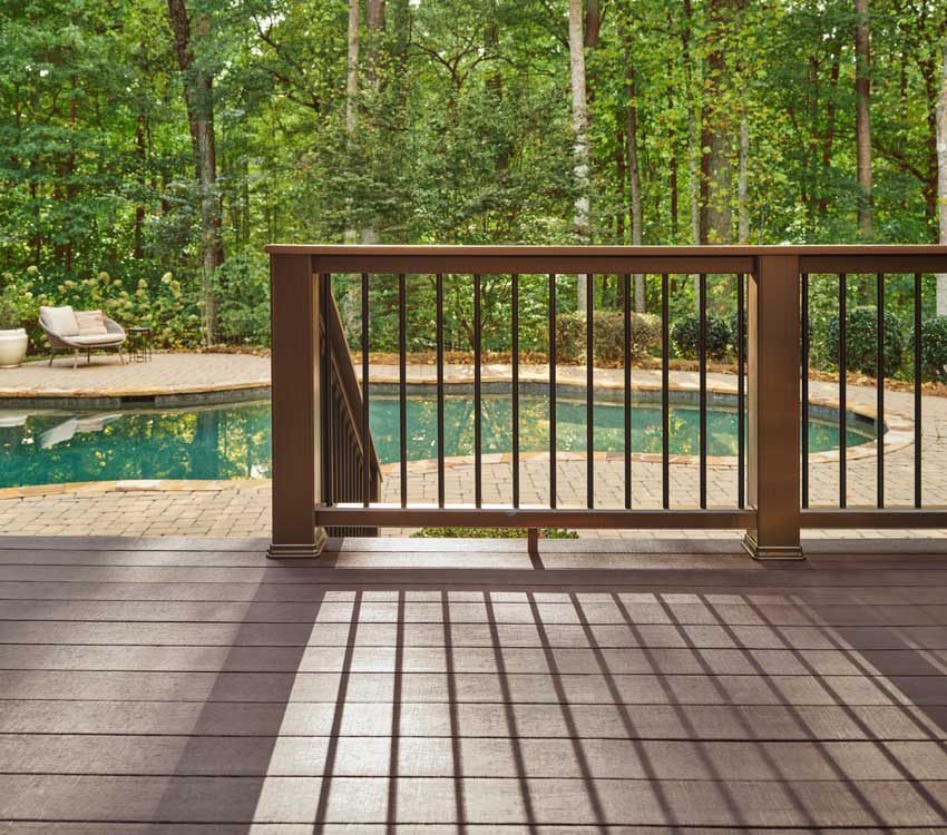 The shadow of a railing stretches across a brown deck while a pool in the background reflects bright sunlight