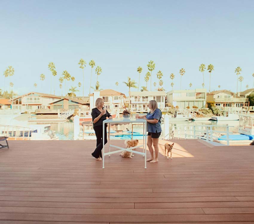 Paddi and a friend stand at a table and chat with a marina and palm trees in the background